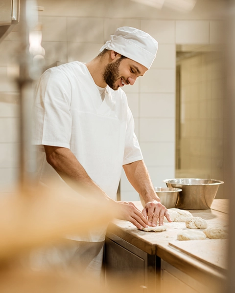 Bäcker in traditioneller Arbeitskleidung formt behutsam Brotteig auf der Arbeitsfläche in der Backstube.